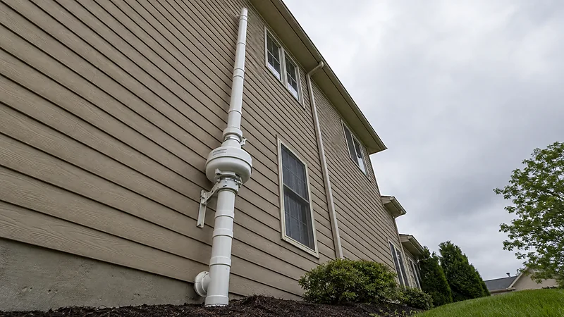 A finished radon vent stack running from grade up an exterior wall past the roofline, with an in-line fan mid-run